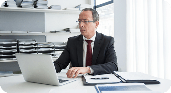 Man in a suit working on a laptop at a desk with documents and a calculator, in an office with shelves of stacked files.