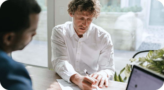 Two men sit at a table; one in a white shirt writes on a document while the other looks on. A laptop is open on the table near them.