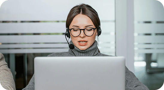 A person wearing glasses and a headset is sitting in front of a laptop, speaking or working in an office setting.