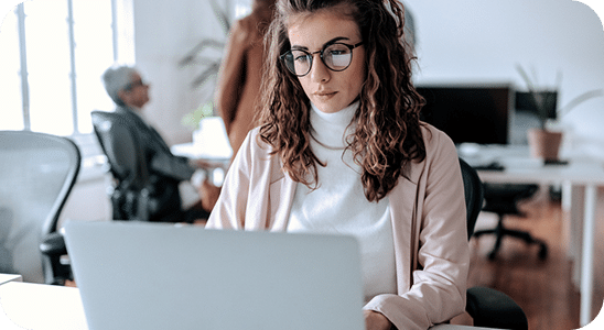 A woman with curly hair and glasses works on a laptop at a desk in a modern office, with other people and office furniture in the background.