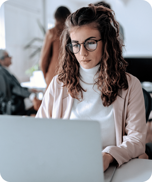 Woman with curly hair and glasses working on a laptop in an office setting with other people in the background.