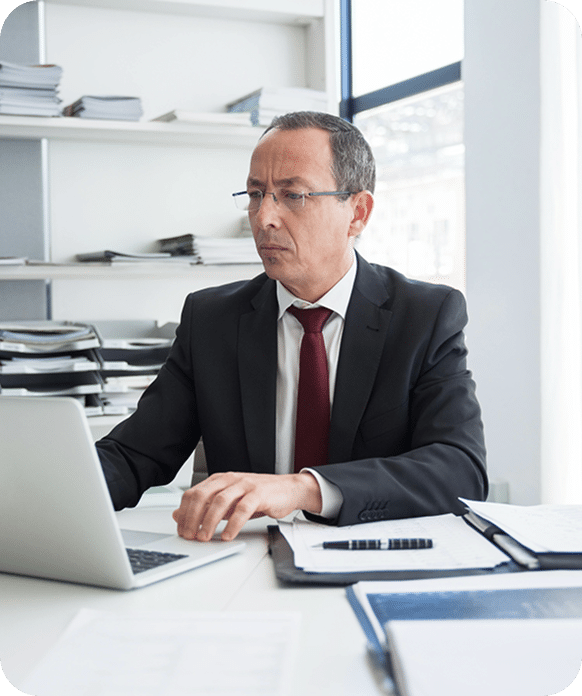 A man in a suit and tie works on a laptop at a desk with documents and files in a bright office setting.