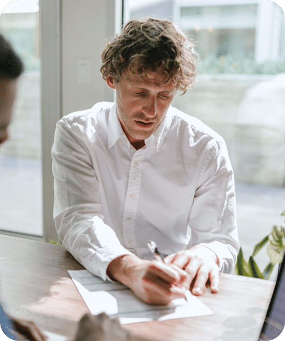 A man in a white shirt sits at a table, filling out a form with a pen in a bright room near a window.