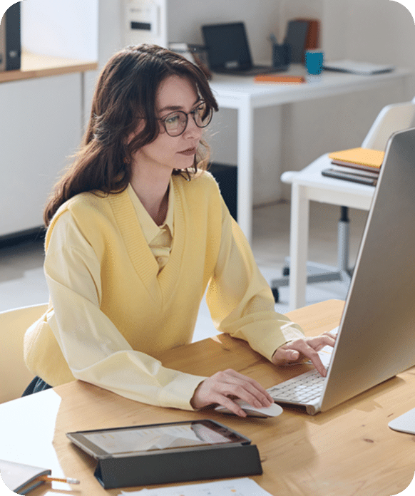 A woman wearing glasses and a yellow vest works on a desktop computer at an office desk, with a tablet and documents nearby.