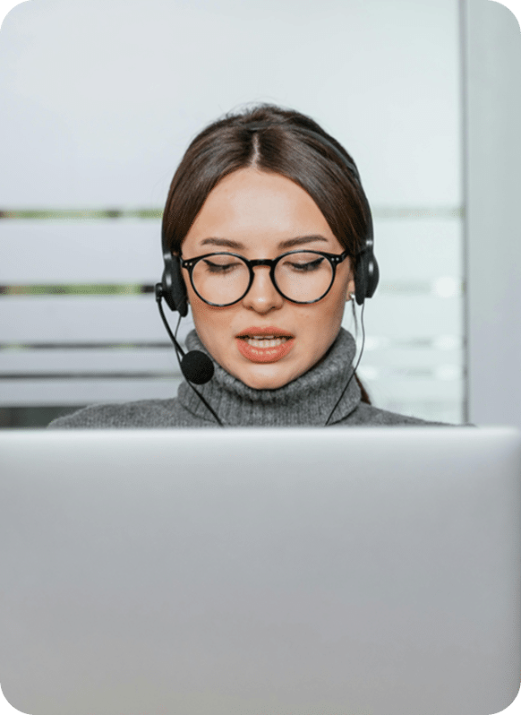 Woman wearing glasses and a headset speaks while looking at a laptop, seated indoors with a blurred background.