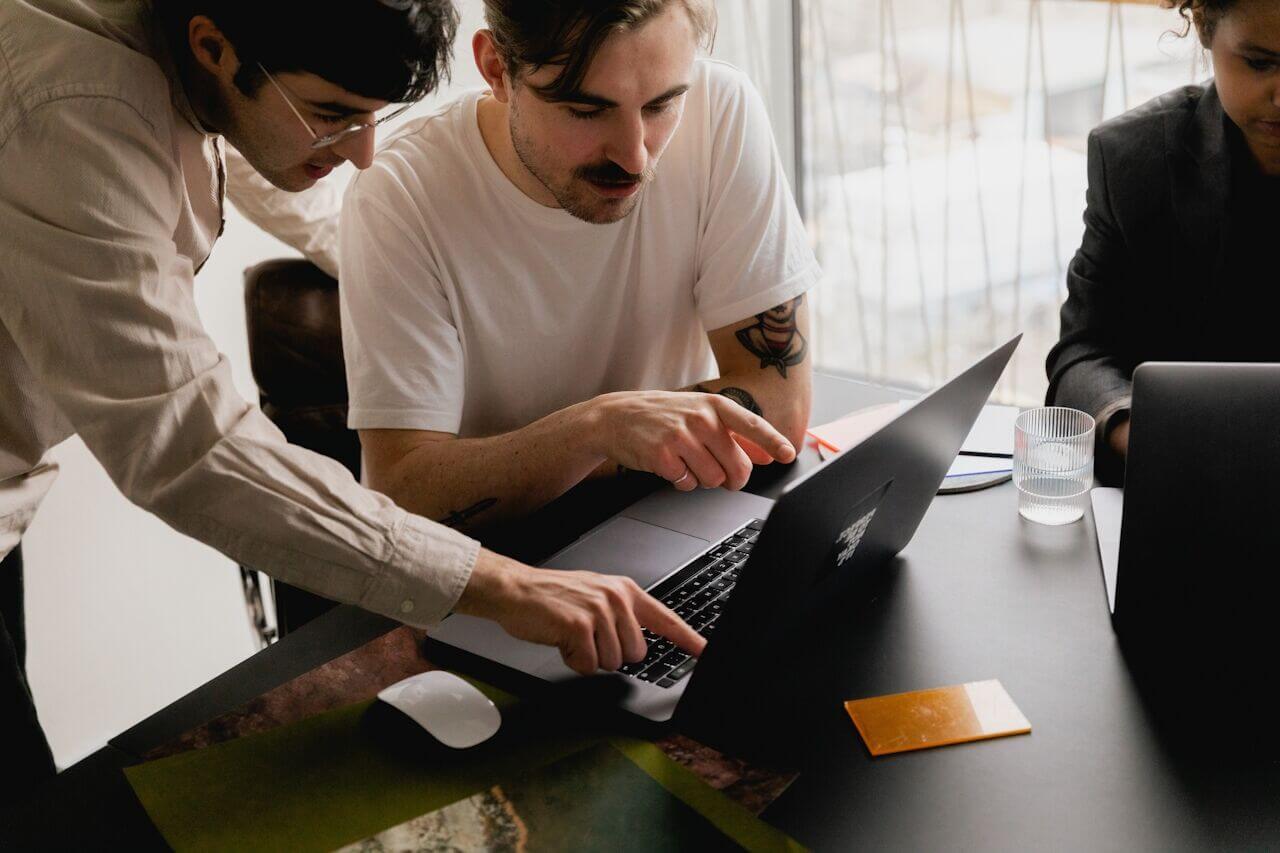 Two people are working together at a desk, looking at a laptop screen and pointing at it, with another person sitting nearby.