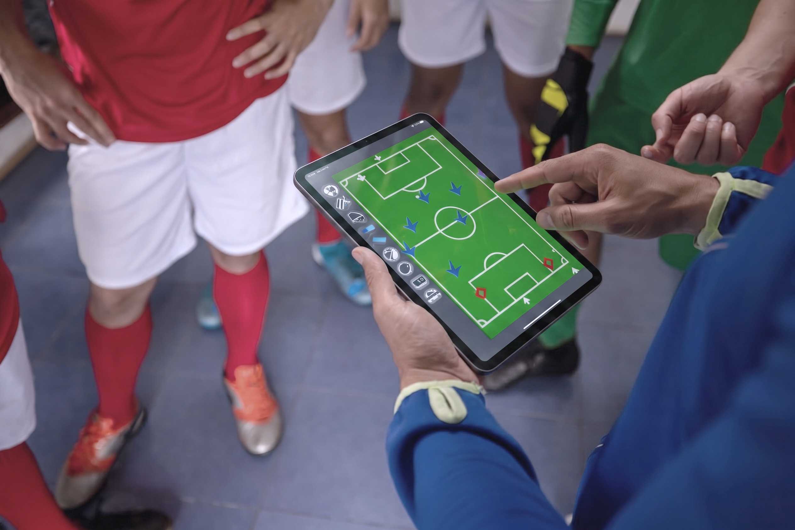 Several soccer players in a locker room stand around a coach holding a tablet that displays a virtual soccer field with tactical positions marked.
