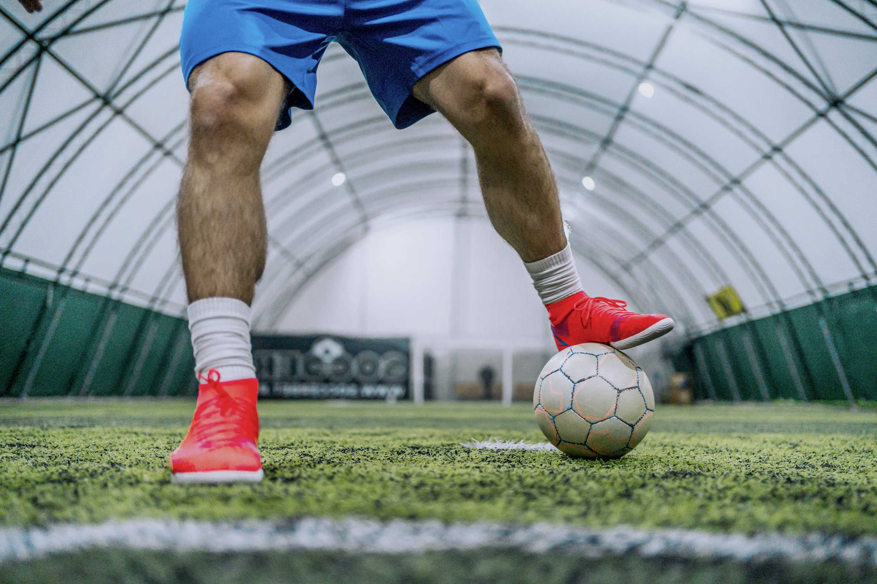 A soccer player in blue shorts and red shoes stands on artificial turf, balancing one foot on a soccer ball inside an indoor sports facility.