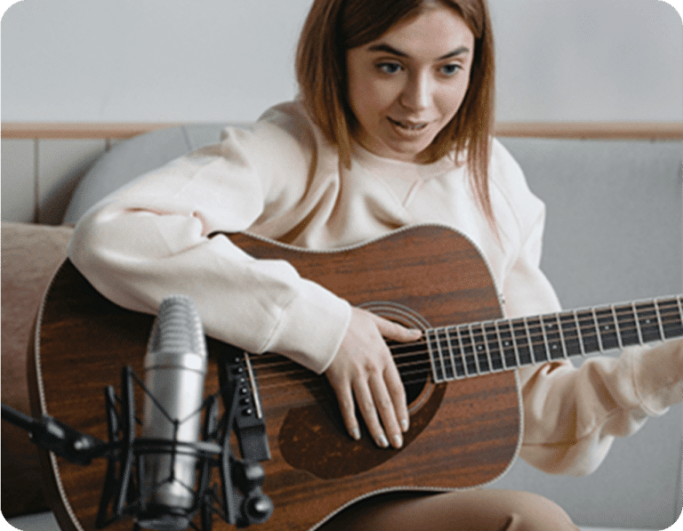 A person playing an acoustic guitar sits in front of a microphone, appearing to record or perform in a home setting—perhaps creating content for a tax guide or resource on global tax compliance for US-based companies.