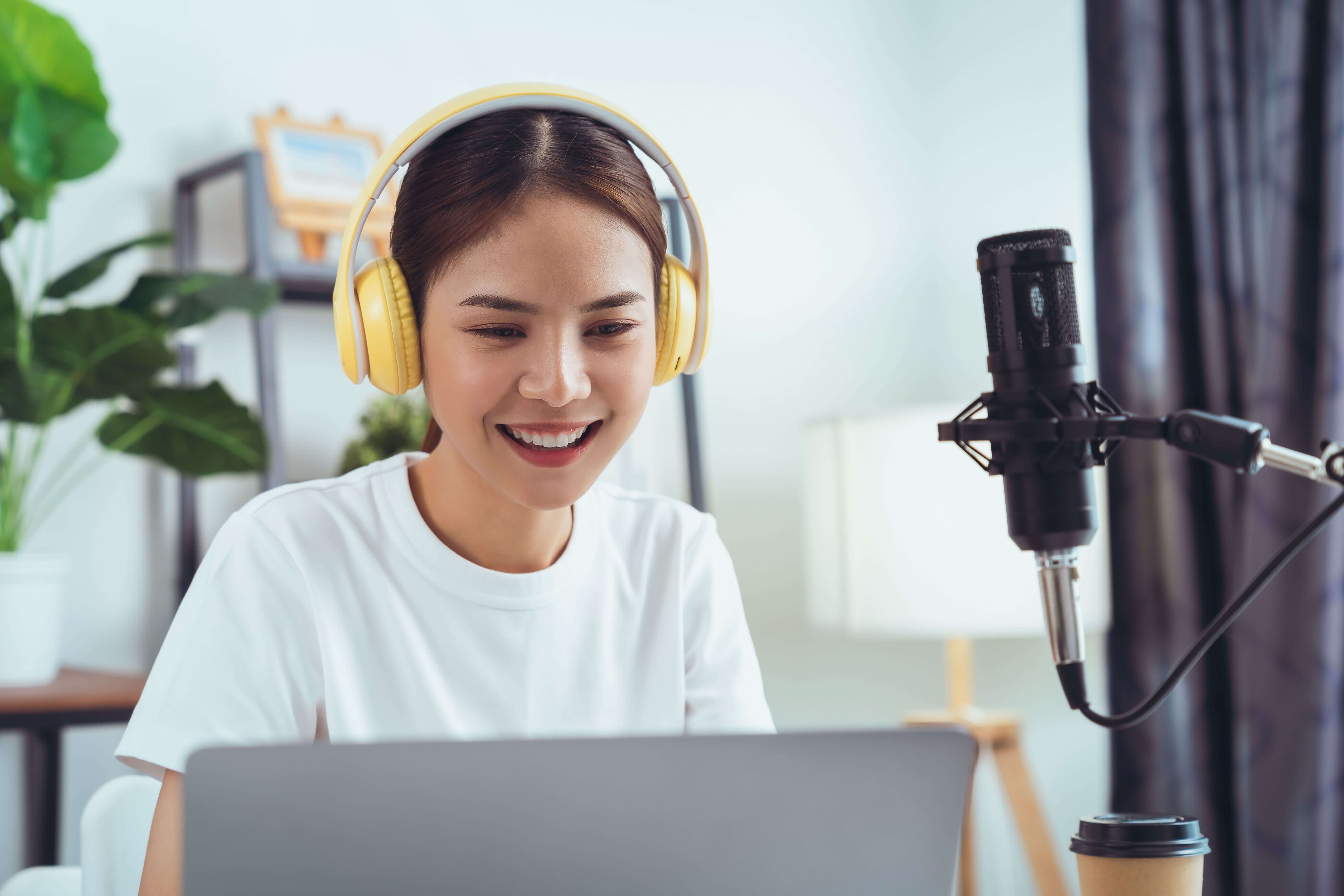 Woman wearing yellow headphones sits at a desk, smiling at her laptop, with a microphone and a coffee cup in front of her in a bright home office setting.