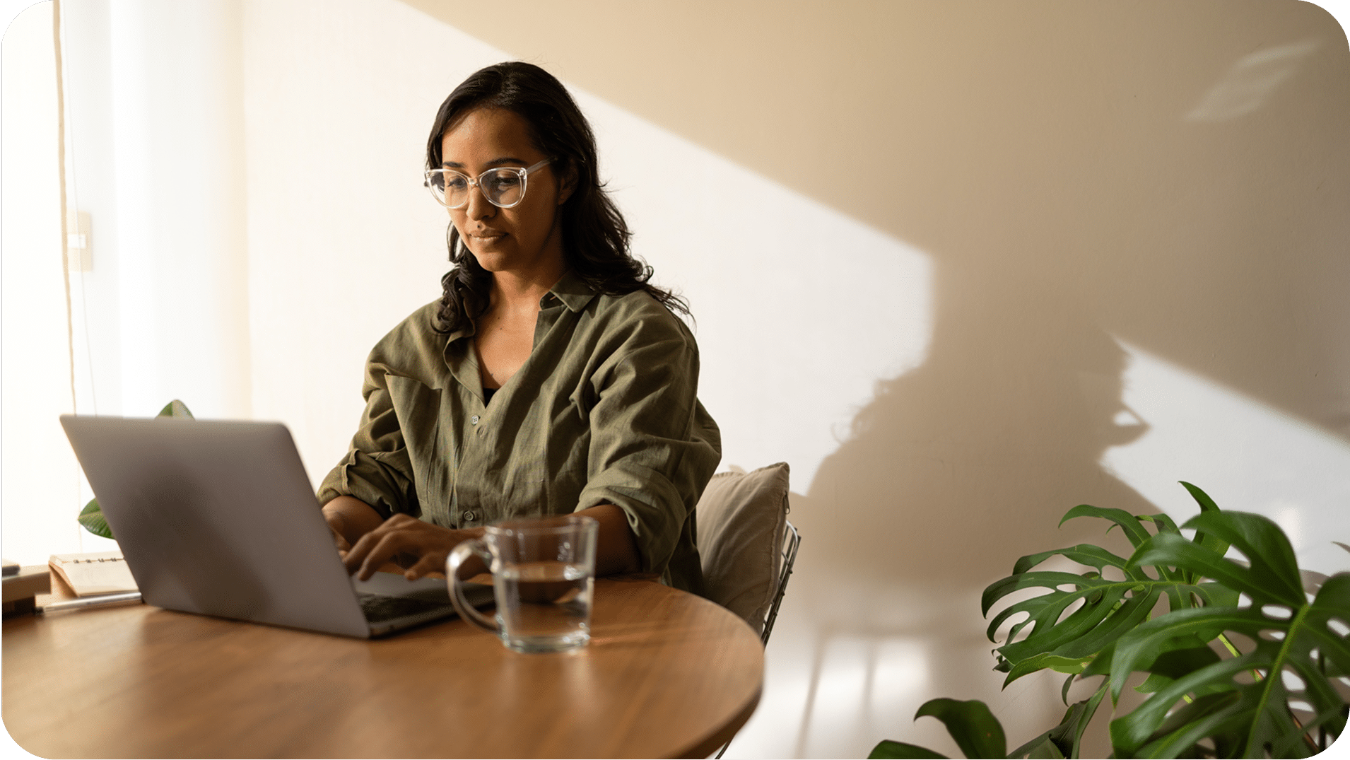 A person wearing glasses works on a laptop at a round wooden table with a glass of water and a potted plant nearby, in a sunlit room.