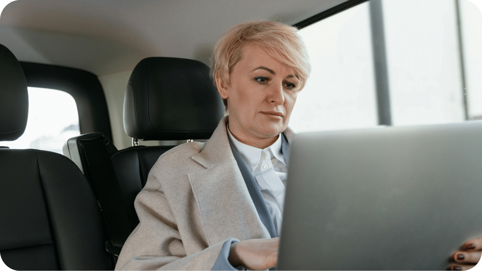 Person with short blond hair uses a laptop while sitting in the back seat of a car, wearing a light-colored coat and white shirt.