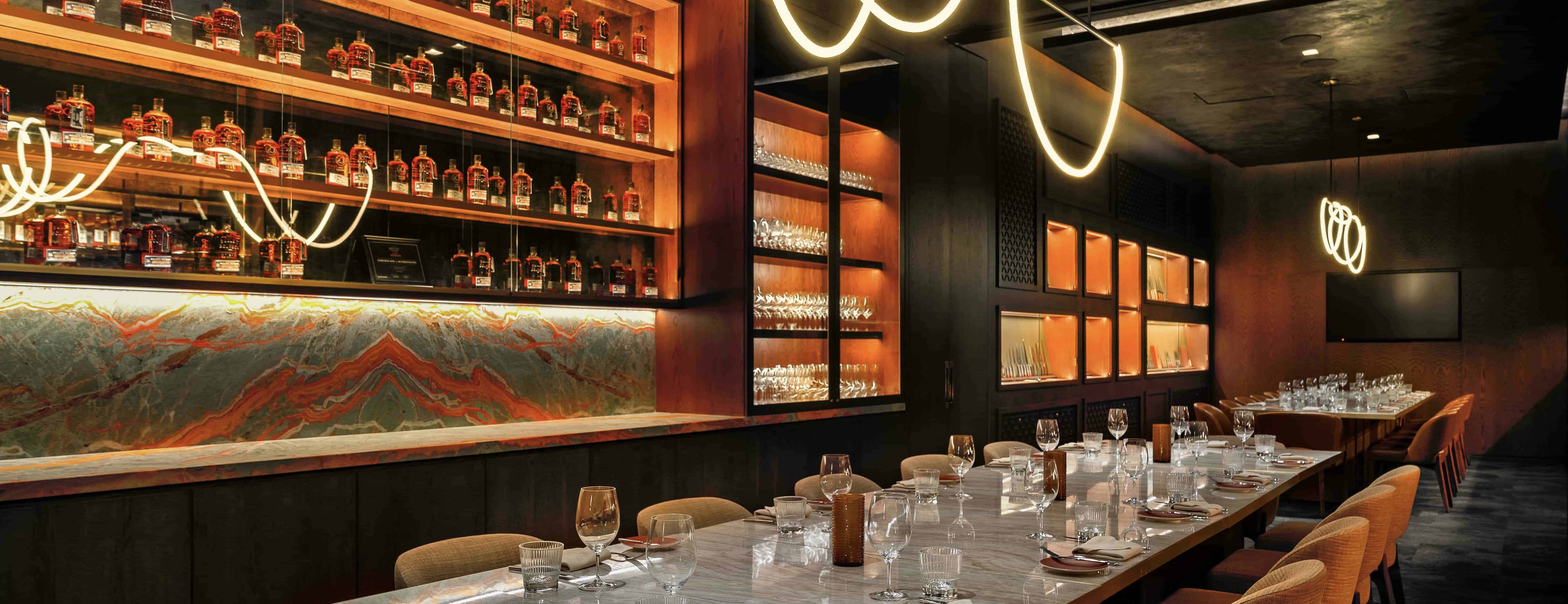 Modern restaurant interior with long dining table, set for guests, shelves of liquor bottles, glassware, warm lighting, and a colorful marble backsplash.