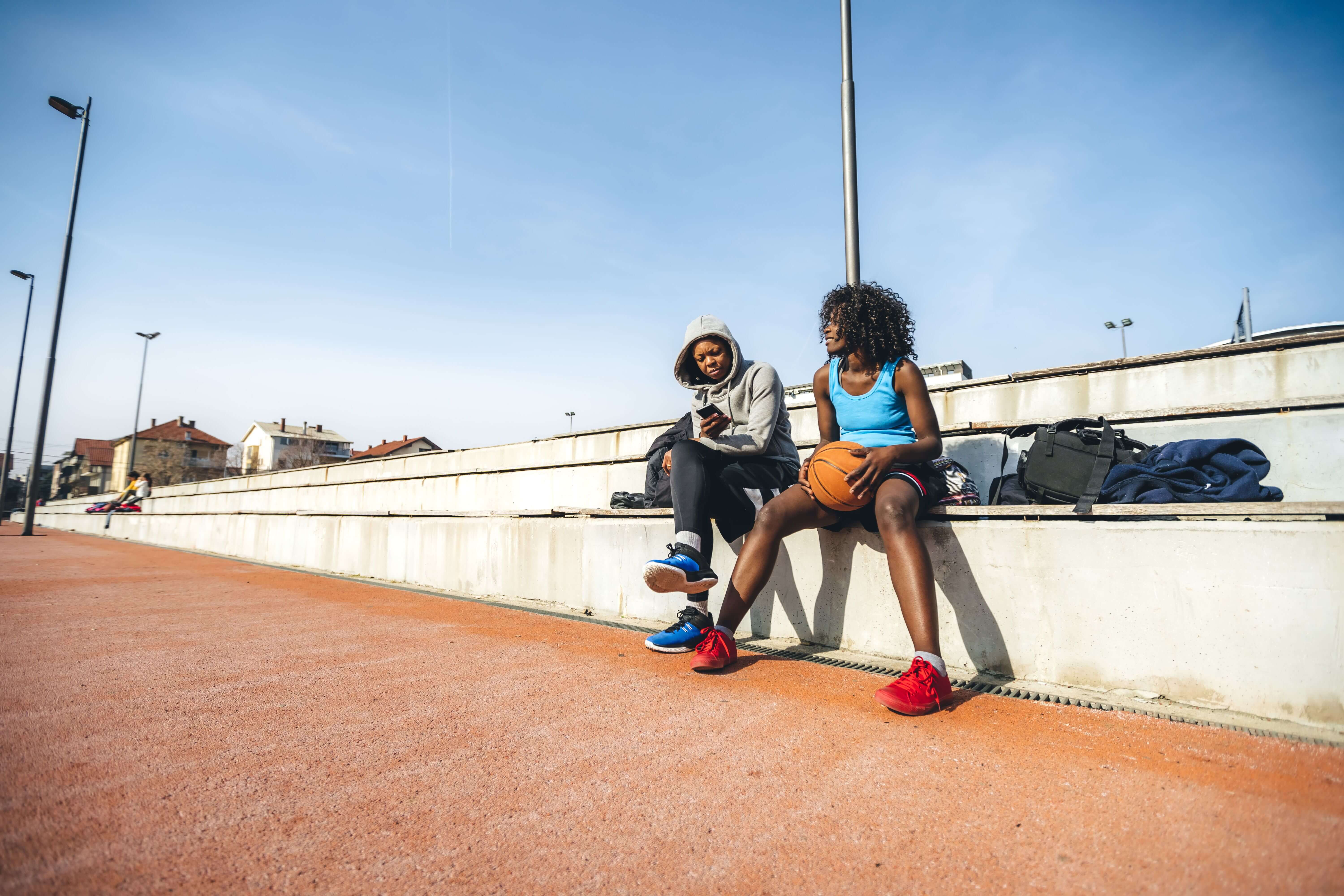 Two people sit on outdoor bleachers; one holds a basketball. They appear to be talking. A clear blue sky and sports bags are visible in the background.