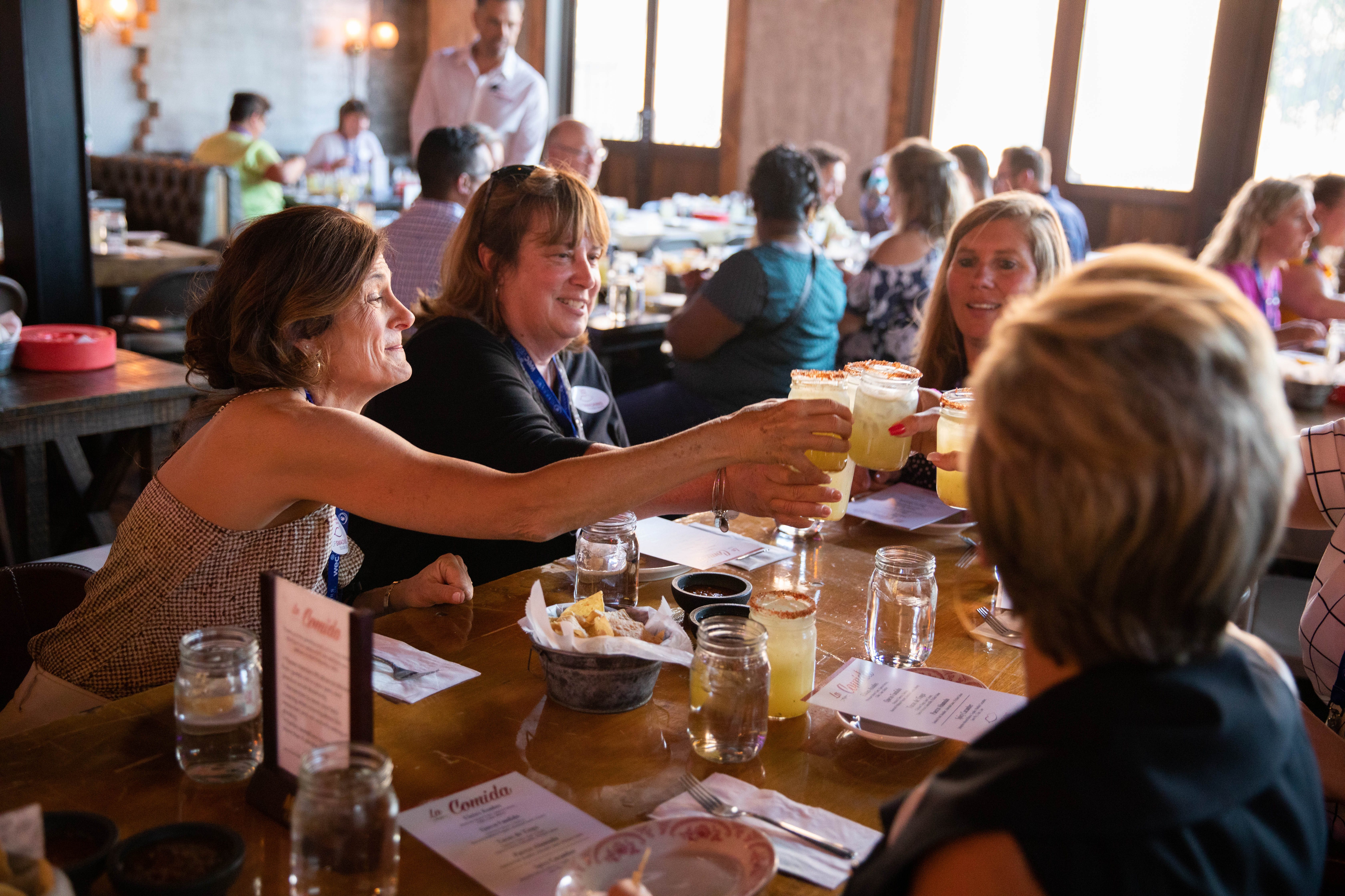 A group of people sit around a restaurant table, raising glasses in a toast. Menus, drinks, and food are visible on the table. The restaurant is busy, with other diners in the background.