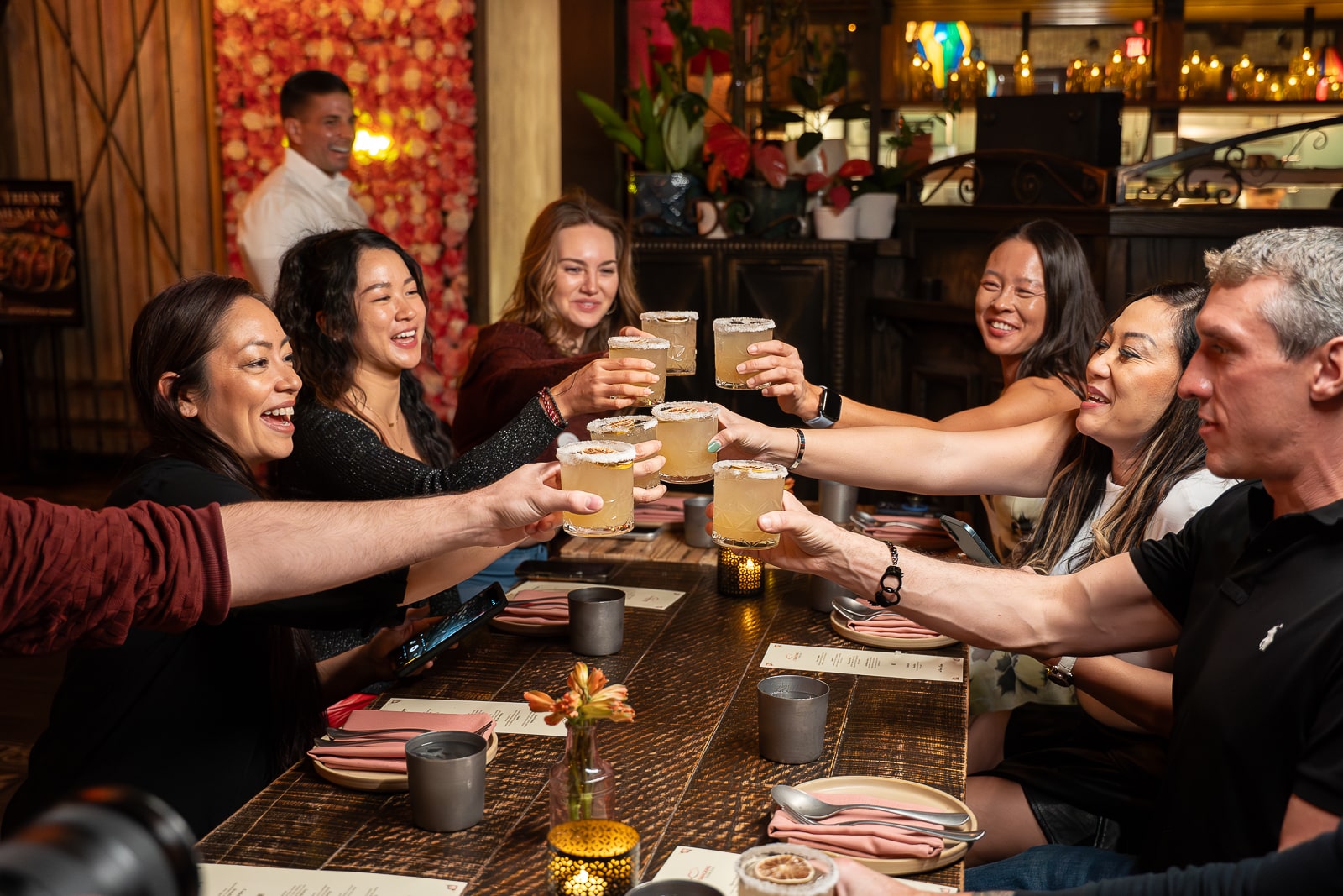 Eight people sitting at a restaurant table raise their drinks together in a toast. Plates, menus, and candles are on the table, and a server stands in the background.