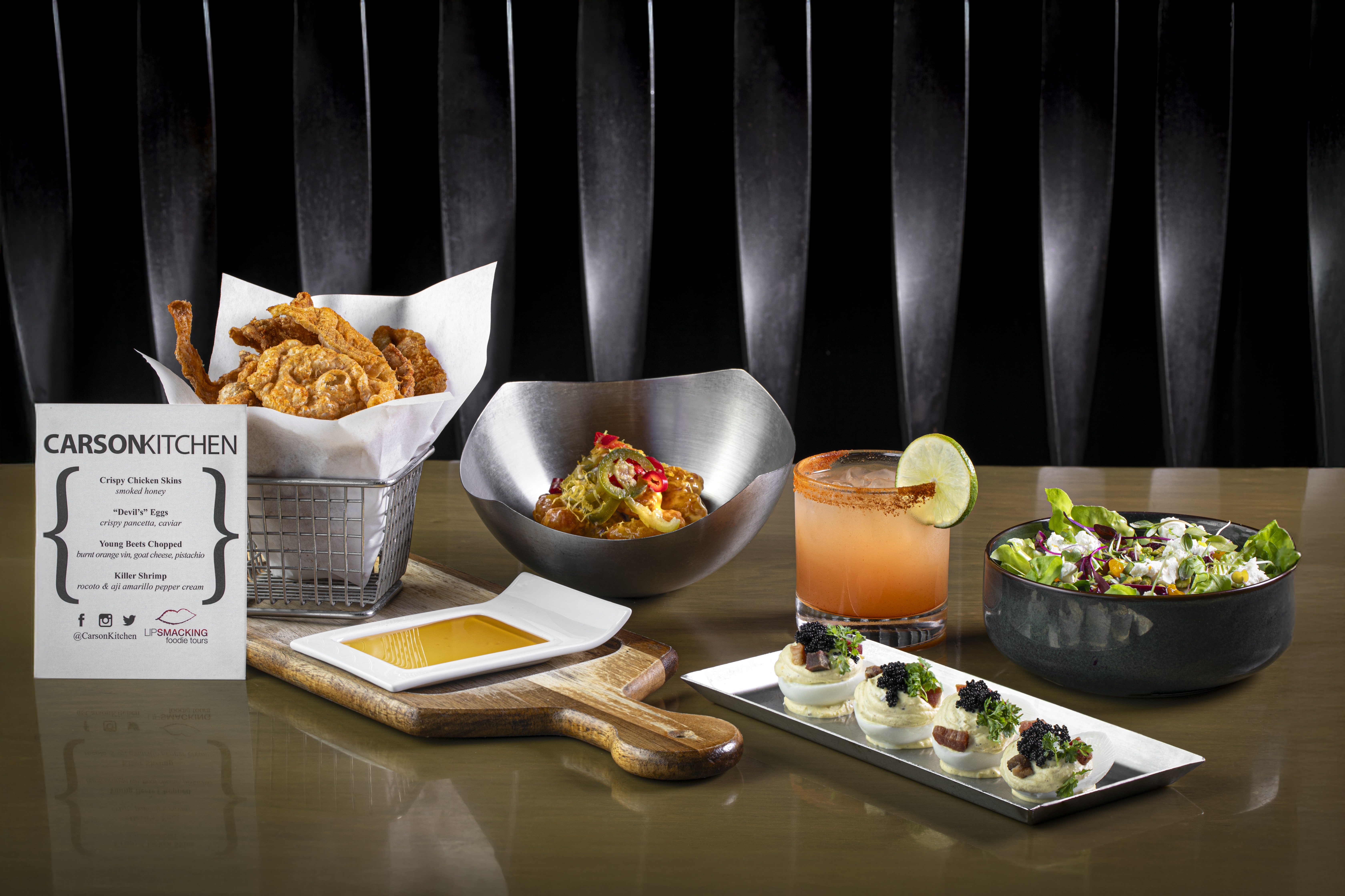 A restaurant table displays crispy chicken skins, pasta, a cocktail, salad, bread with dip, deviled eggs with caviar, and a Carson Kitchen menu on a glossy surface with a black background.