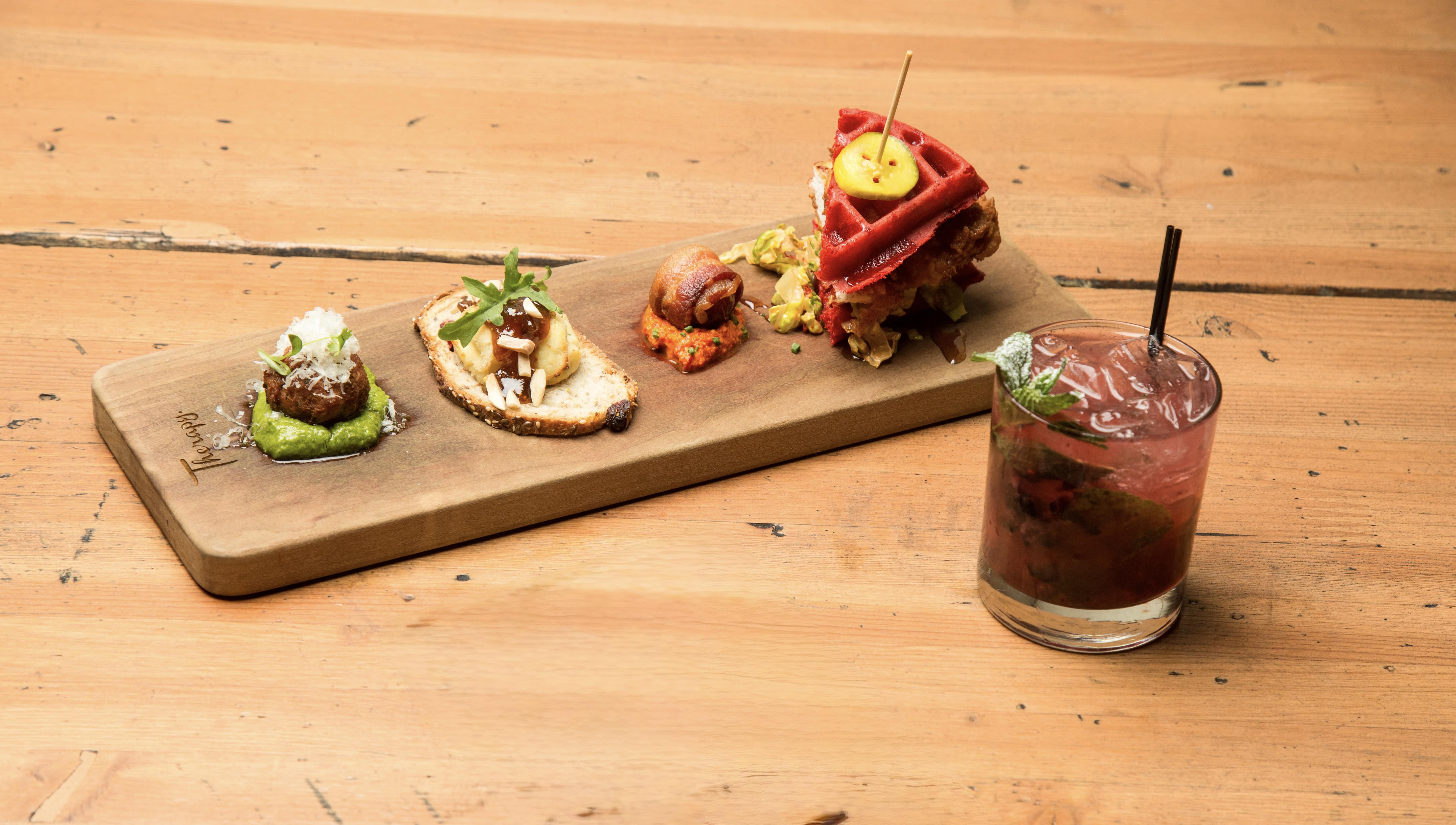 A wooden board with three appetizers and a sandwich, next to a glass of pink cocktail with ice, mint, and a straw on a wooden table.