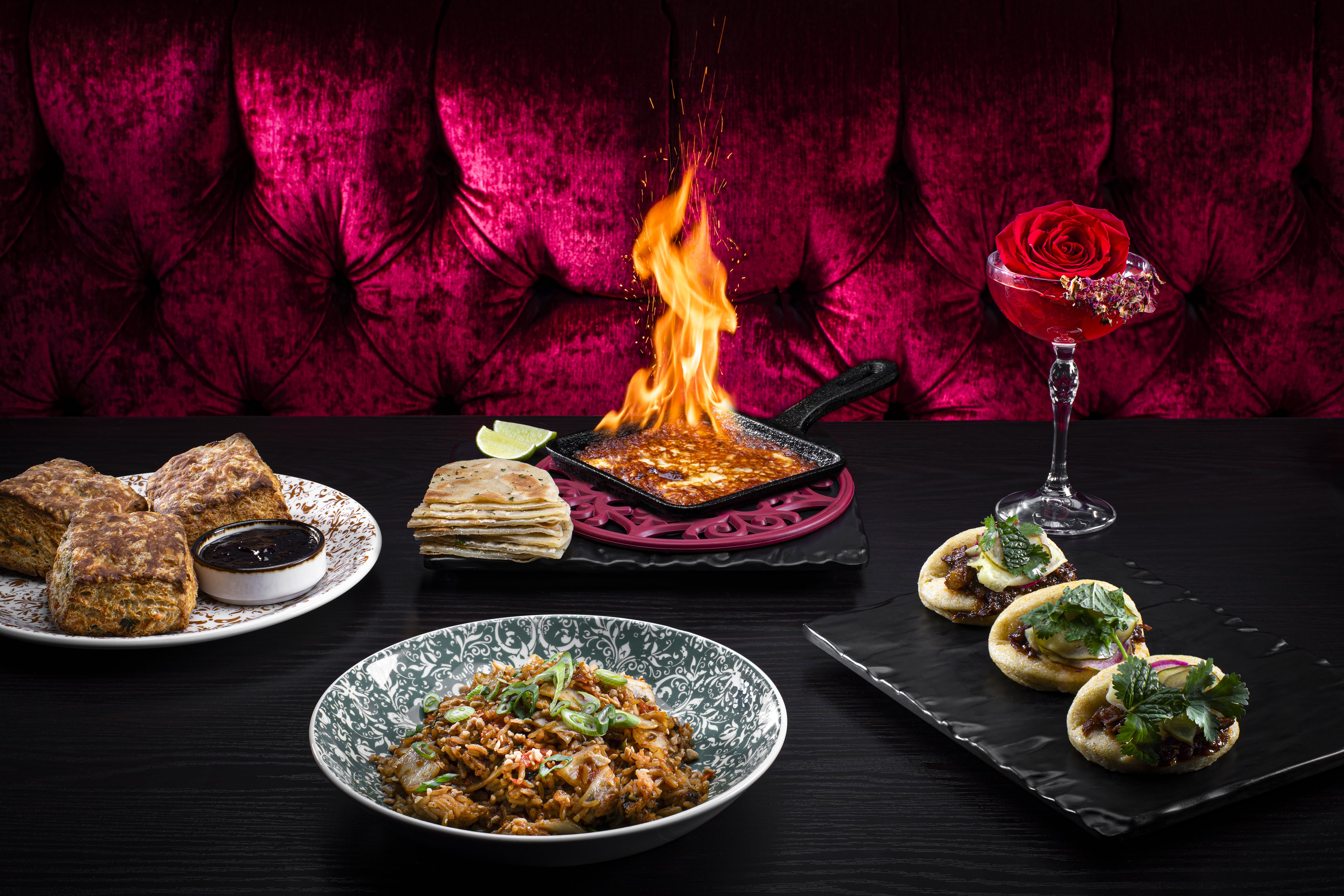 An elegant table with scones, flatbread, a fiery cheese dip, a cocktail with a rose, mini tacos, and a pasta dish, set against a red velvet backdrop.
