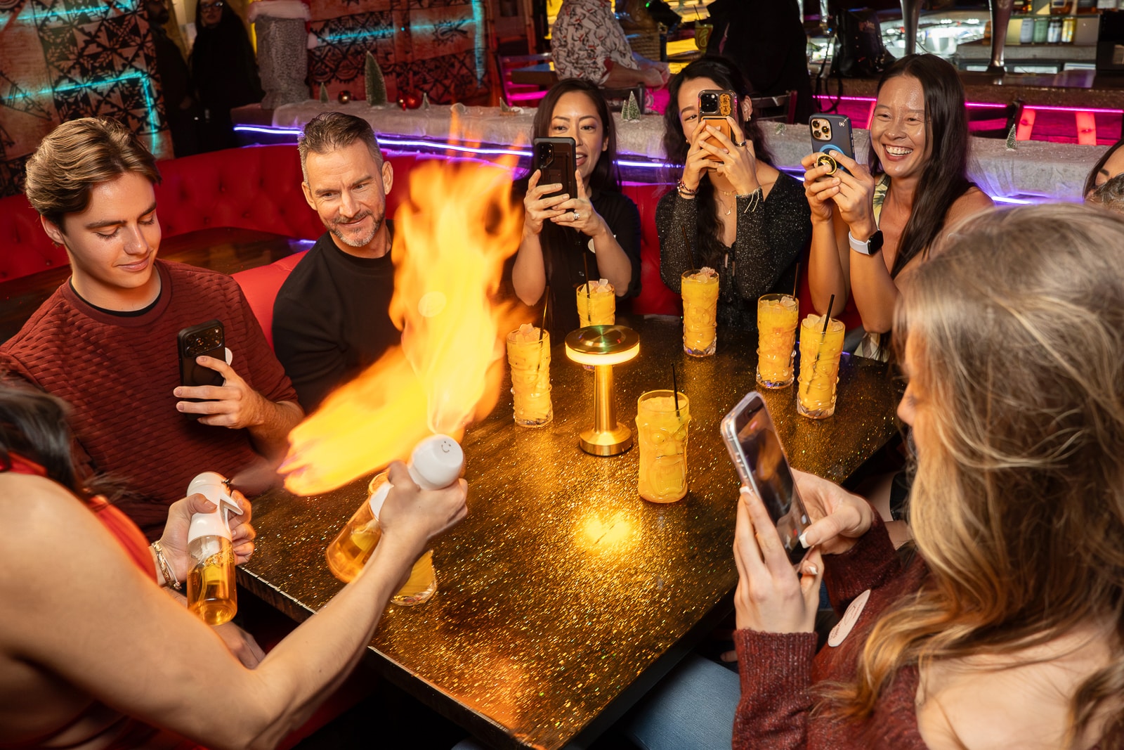 A group of people sit around a table with drinks, taking photos as one person creates a flame with a lighter and a bottle.