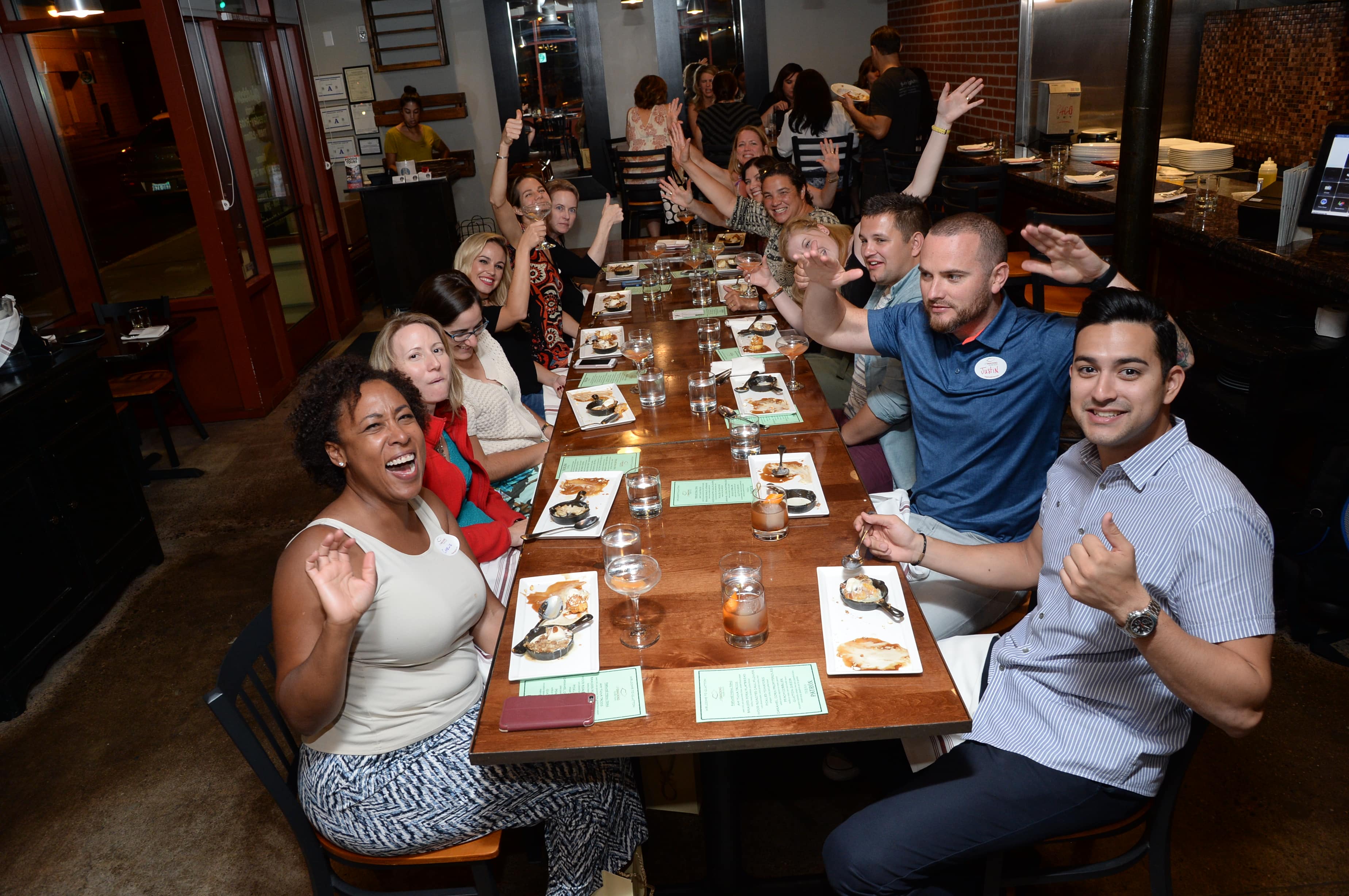 A group of people sit around a long restaurant table, smiling and waving at the camera, with plates of food and drinks in front of them.