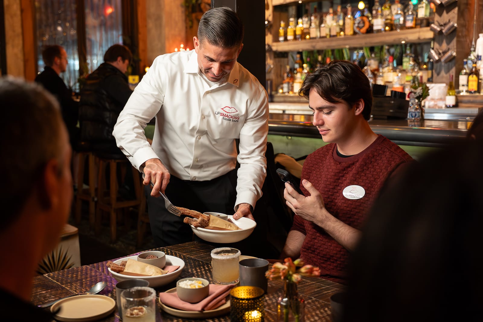 A waiter serves food to a seated man looking at his phone in a restaurant with drinks and dishes on the table.
