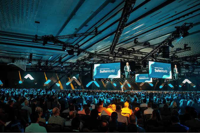A large conference hall filled with attendees faces a stage where a speaker presents at SuiteWorld, with multiple screens displaying the event name.