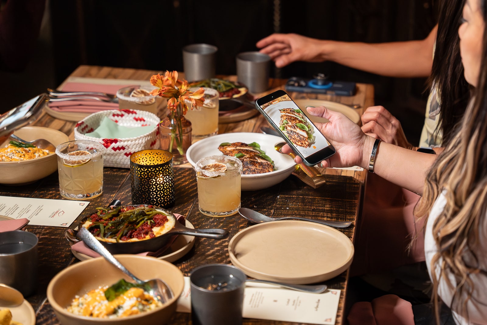 A person photographs a dish of grilled chicken with a smartphone at a dinner table filled with various bowls, plates, cocktails, and a candle.