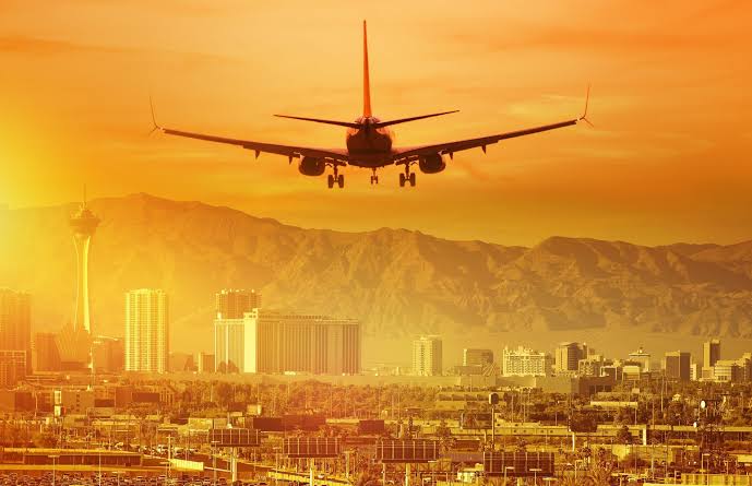An airplane flies over a cityscape with mountains in the background at sunset, with tall buildings and a warm orange sky visible.