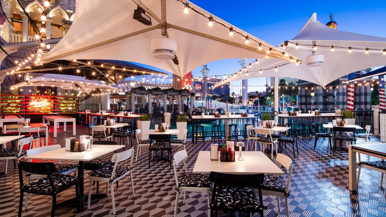 Outdoor restaurant patio with empty tables and chairs, surrounded by string lights and large white canopy, during dusk. Patterned floor tiles and cityscape visible in the background.