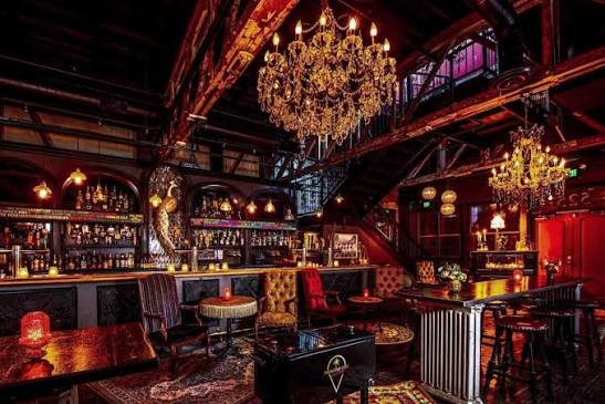 Dimly lit bar with chandeliers, a mezzanine level, vintage furniture, ornate decor, and a fully stocked bar in the background.