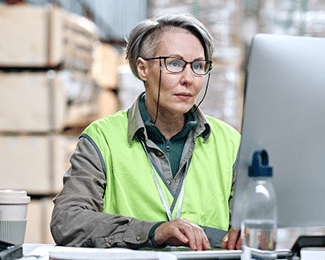 A person with short grey hair wearing glasses and a lime green safety vest sits at a desk using a computer in a warehouse setting.