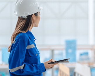 A woman in a hard hat and blue work uniform holds a tablet in an industrial facility.